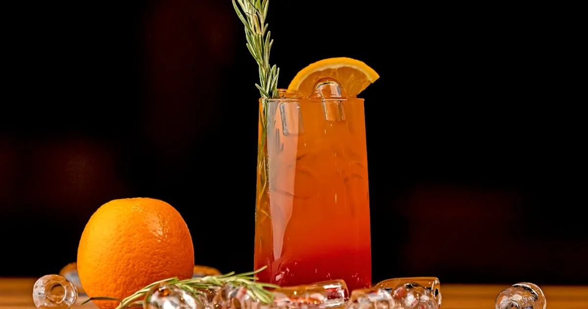 Tall mocktail glass with orange slice and rosemary garnish, ice and a whole orange on a bar counter