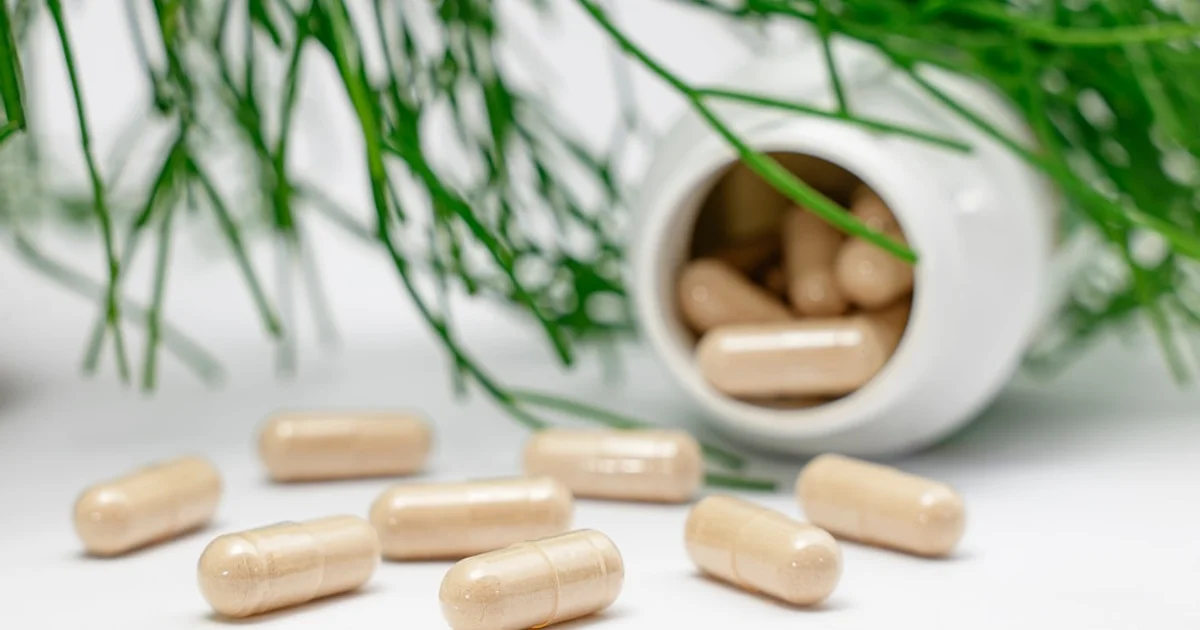 Tan supplement capsules spilled from an unlabeled white bottle next to a green plant