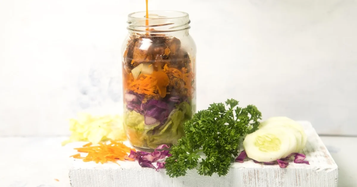 Dressing being poured into a layered mason jar salad with cabbage, carrot, and cucumber